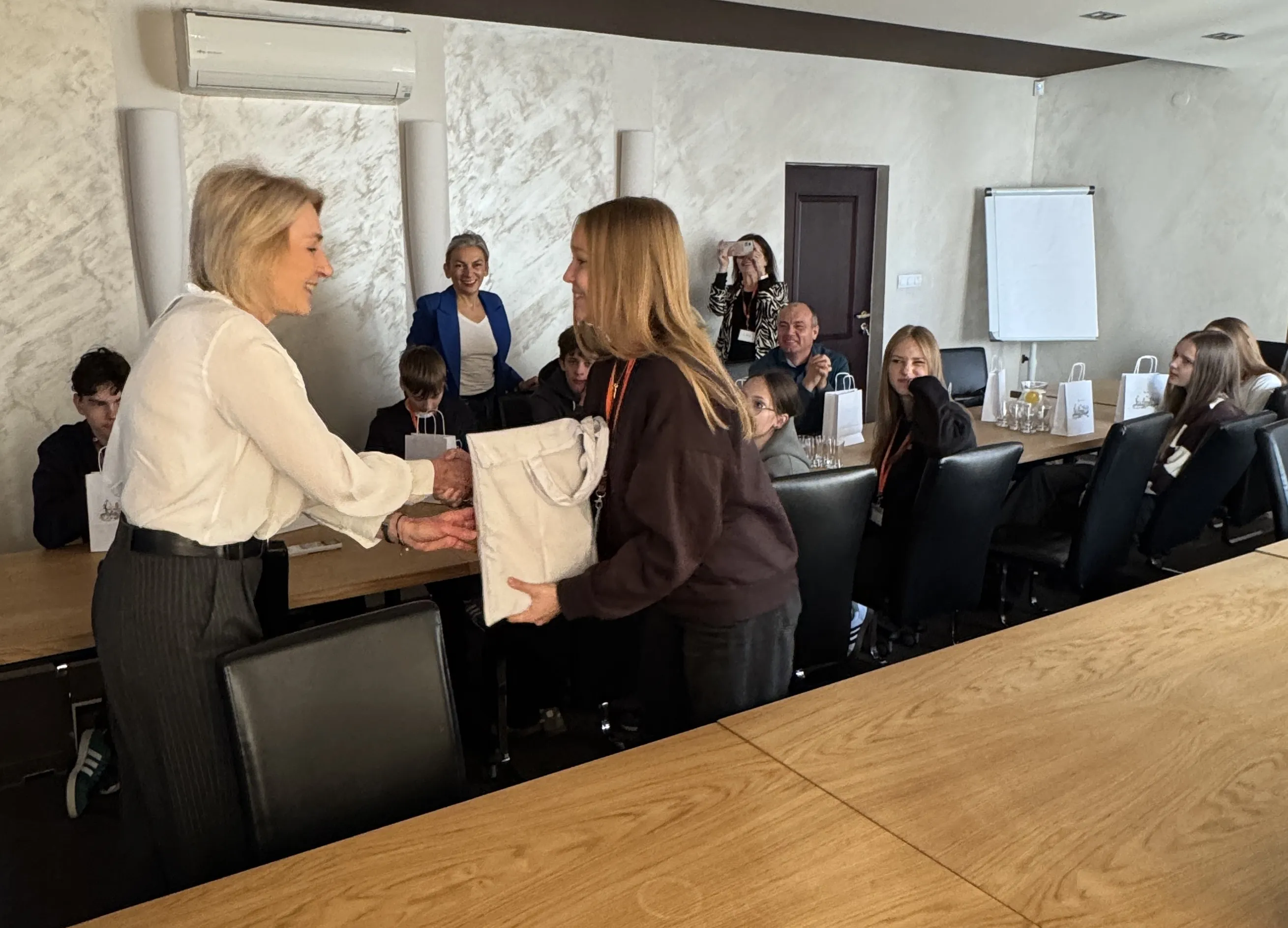 Kobieta wręcza upominek młodej osobie w sali konferencyjnej, wokół stołu siedzi grupa ludzi obserwujących tę scenę, jedna osoba robi zdjęcie telefonem, na stole stoją białe torby i szklanki z napojami.